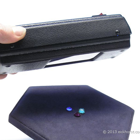 Hand holding a UV lamp over gemstones on a dark pad, showing fluorescence under ultraviolet light during gem inspection.
